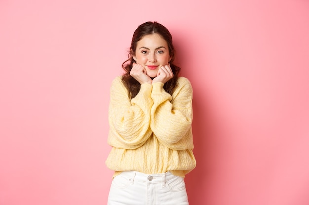 Cute silly girl holding hands on jawline and watching something happening, look with temptation and love at camera, smiling happy, standing pink wall.