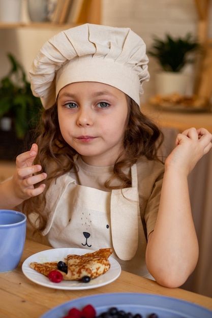 シェフの帽子とエプロンでかわいい6歳の女の子がパンケーキを食べて木製のテーブルに座っています