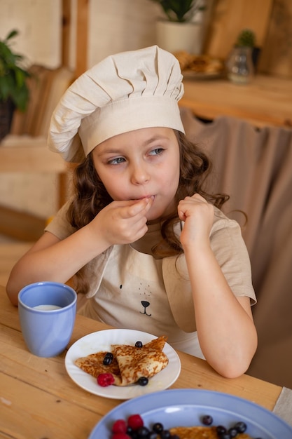 シェフの帽子とエプロンでかわいい6歳の女の子がパンケーキを食べて木製のテーブルに座っています