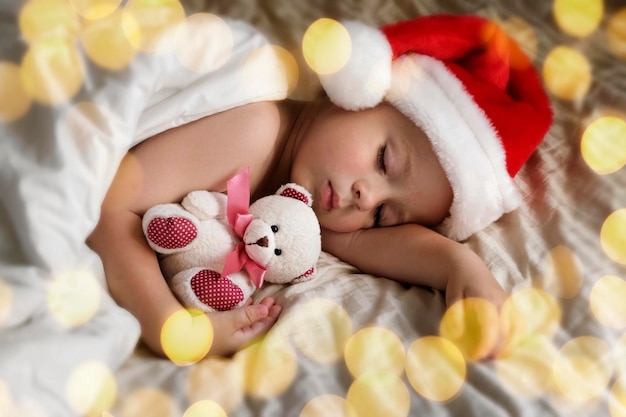 サンタ クロースの帽子をかぶったかわいい眠っている若い男の子が家でベッドに横たわっています クリスマス ライト 冬の休日の概念 夢 幼児 赤ちゃん クリスマス メリー クリスマス 新年あけましておめでとうございます 面白い小さな子供