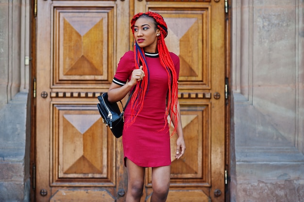 Cute and slim woman in red dress with dreadlocks and backpack posed against school large wooden door