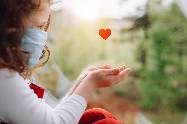 Cute small girl wear face blue mask sitting on sill near little heart as a way to show thank your nurses thanking doctors and medical staff working in hospitals during coronavirus COVID-19 pandemics