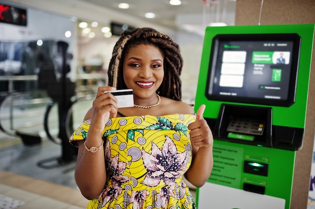 Cute small height african american girl with dreadlocks, wear at coloured yellow dress, against ATM with credit card at hand.