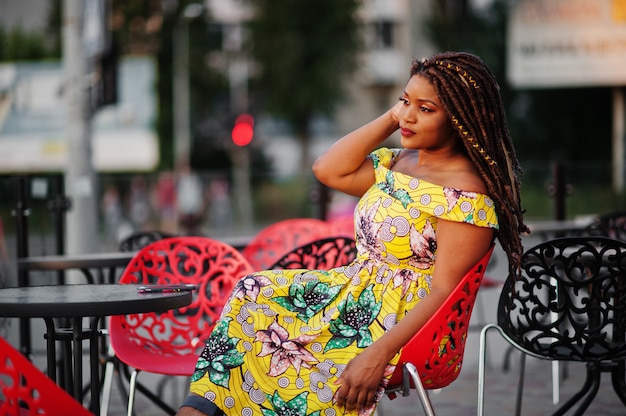 Cute small height woman with dreadlocks, wear at coloured yellow dress, sitting at outdoor cafe on red chair