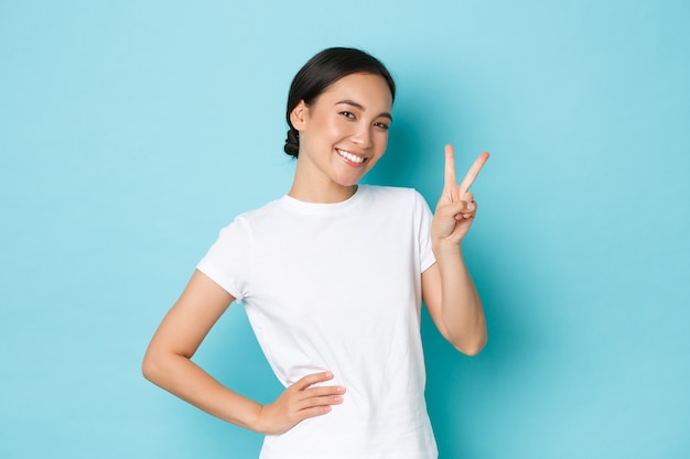 Cute smiling asian female in white t-shirt, showing peace kawaii sign and looking adorable, flirting, staying positive or optimistic, standing blue wall happy attitude.