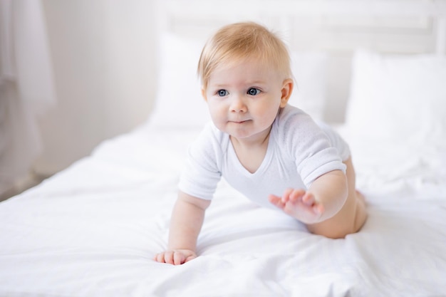 귀여운 웃는 아기는 집에 있는 흰색 침대에서 기어가는 법을 배웁니다. 작은 금발의 아기가 아침에 일어났습니다. 행복한 어린 시절과 가족