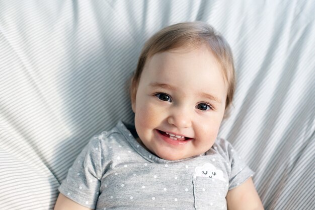 Cute smiling baby girl laying in the bed in morning sun light. Grey colors