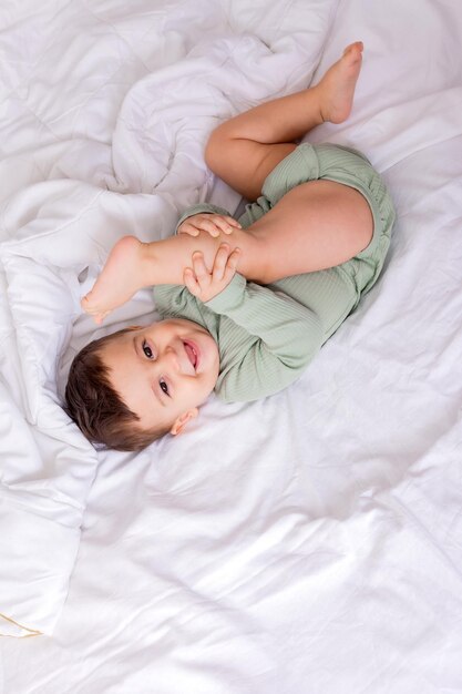 Cute smiling baby in a green cotton bodysuit is lying on a white bed on his back top view high