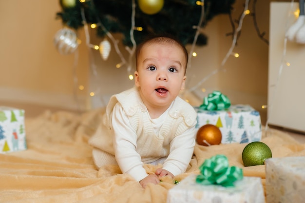 Cute smiling baby is lying under a festive Christmas tree and playing with gifts. Christmas and New Year celebrations.
