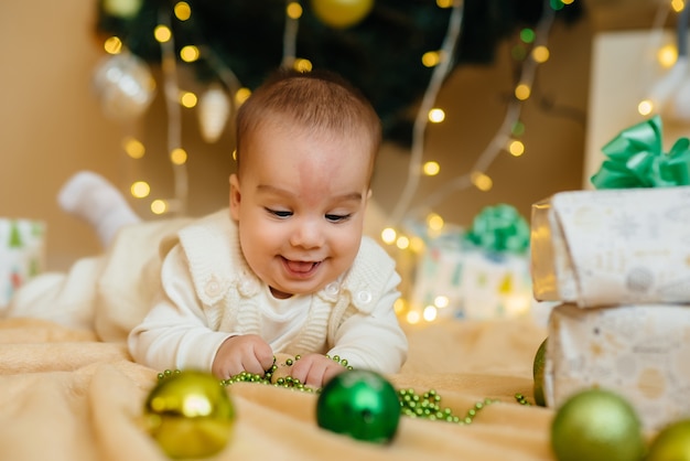 Cute smiling baby is lying under a festive Christmas tree and playing with gifts 