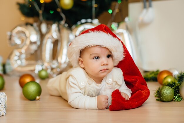 Cute smiling baby is lying under a festive Christmas tree and playing with gifts 