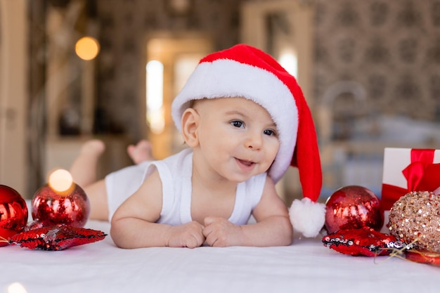 Cute smiling baby in a white bodysuit and Santa hat is lying on his stomach on a white sheet