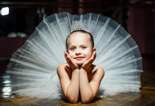 Photo cute smiling ballerina kid in a white splendid tutu laying on the wooden floor and posing to the camera child is grinning and resting her head in her hands