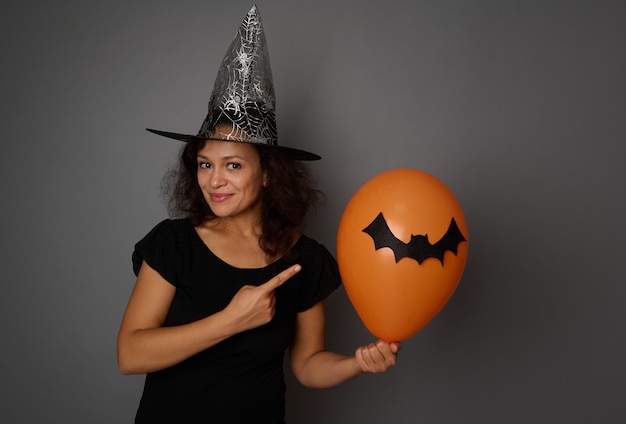 Cute smiling beautiful and joyful woman dressed in Halloween carnival black witch clothes, points on an orange air ball with a handmade bat, standing isolated over gray background with copy space