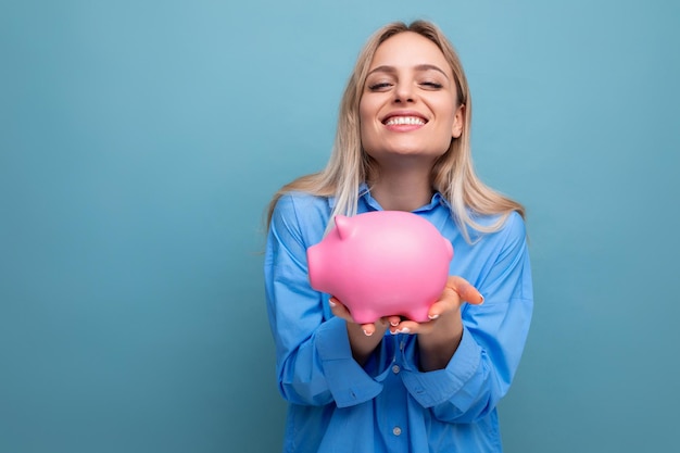 Cute smiling blond young adult woman holding out her piggy bank where she keeps savings on a blue