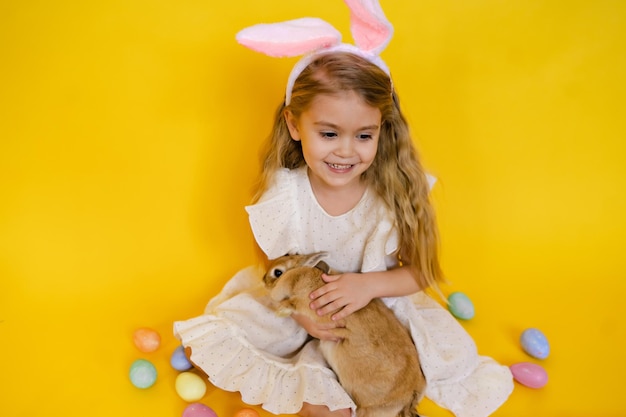 Cute smiling blonde woman with bunny ears holding a red rabbit in her hands on a yellow studio