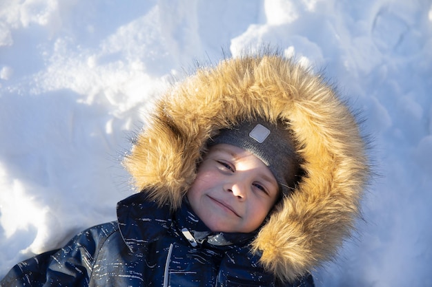 帽子と毛皮のフードでかわいい笑顔の男の子は、彼が楽しんでいる雪の中に横たわっています冬の活動