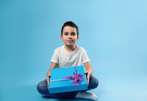 Cute smiling boy posing with a birthday gift in his hands on blue surface with copy space