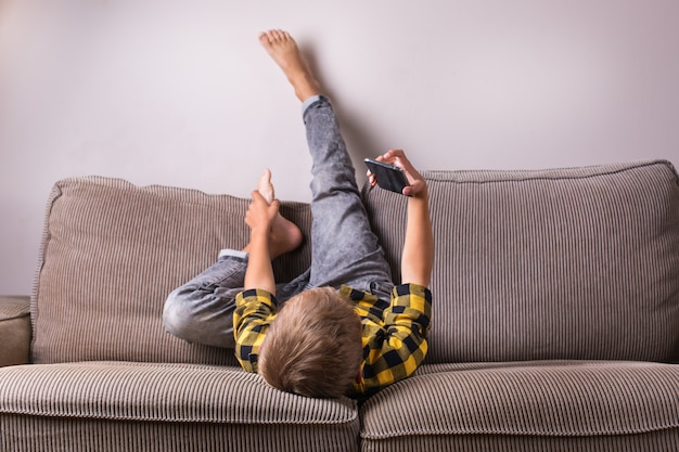 Cute smiling boy talking with family and friends, making video call on the phone, sitting on a couch. Stay at home, lockdown, social distancing, quarantine.