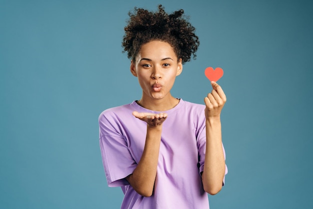 Cute smiling brunette woman holding pink heart and sending air kiss to the camera while supporting another women. International breast cancer awareness. Indoor studio shot isolated on blue background