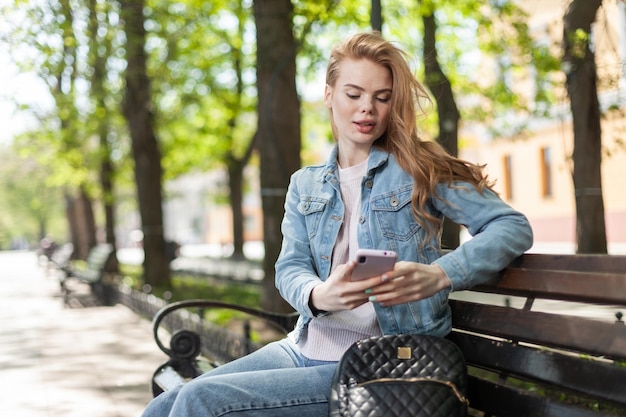 かわいい笑顔の白人女性が街のベンチに座り、スマートフォンを覗き込む