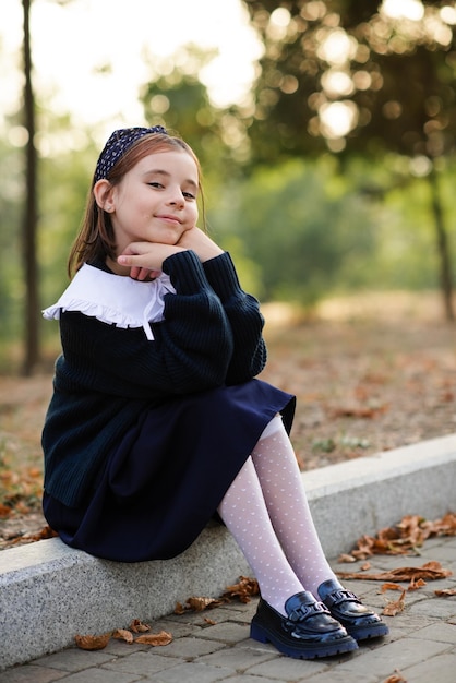 Cute smiling child pupil girl 6-7 year old wear school uniform posing in patk outdoor Look at camera