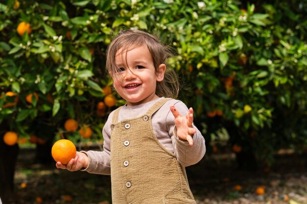 Cute smiling child with fresh orange looking at camera against tropical tree in summer