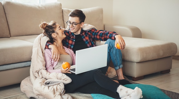 Coppie sorridenti sveglie che abbracciano sul pavimento vicino al divano coperto con un copriletto utilizzando un computer portatile e tenendo le arance