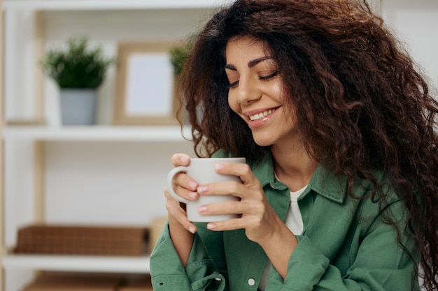 Cute smiling enjoyed pretty tanned curly latin lady look with\
love aside drink tea coffee in casual shirt sit on chair in home\
modern interior background copy space banner nostalgia concept