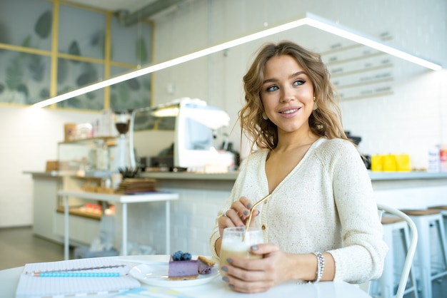 Cute smiling female in casualawear sitting by table in cafe, having cappuccino and tasty bluberry cake at break