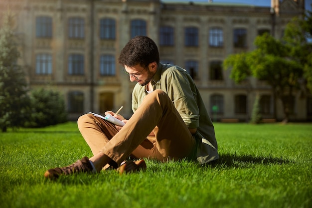 Cute smiling focused undergraduate in round eyeglasses and casual clothes sitting cross-legged on the grass