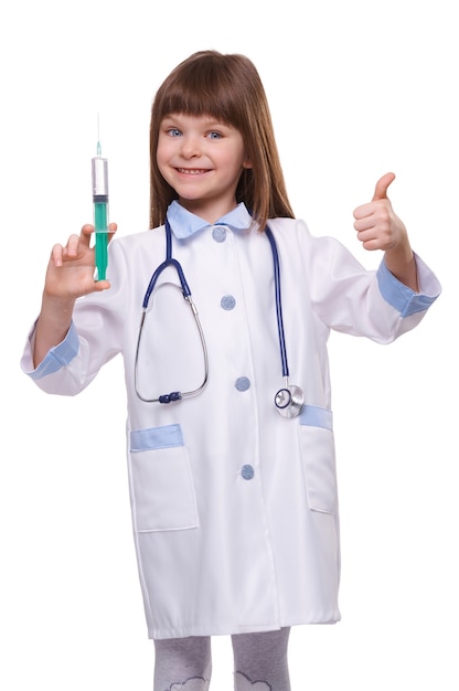 Medico sorridente sveglio della ragazza in siringa della tenuta dell'abito medico e che mostra il pollice su su fondo isolato bianco