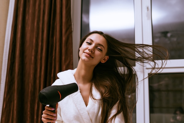 Cute smiling girl dries her long hair with a hairdryer