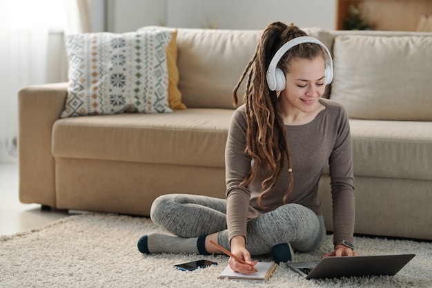 Ragazza sorridente sveglia con le cuffie che prendono appunti mentre sedendosi sul pavimento dal divano davanti al computer portatile