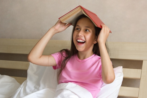 Carina ragazza sorridente con lunghi capelli scuri si siede a letto durante il giorno tiene il tetto del libro sopra la sua testa