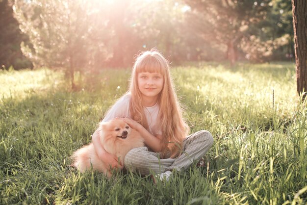 Cute smiling kid girl having fun with domestic pet dog sitting on green grass