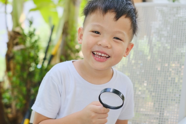 Cute smiling kindergarten boy exploring environment by looking through a magnifying glass in garden