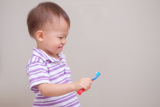 Cute smiling little asian toddler boy child wearing purple shirt holding toothbrush and learn to brushing teeth in bathroom at home