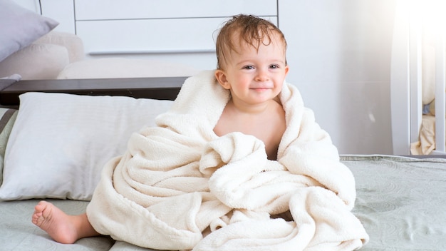 Cute smiling little boy covered in white towel after bath sitting on bed.