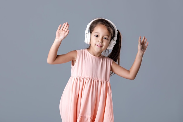 Cute smiling little child girl in pink dress and headphones listens to music and dances on gray background.