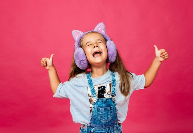 Cute smiling little girl having fun in fur headphones on a pink background.