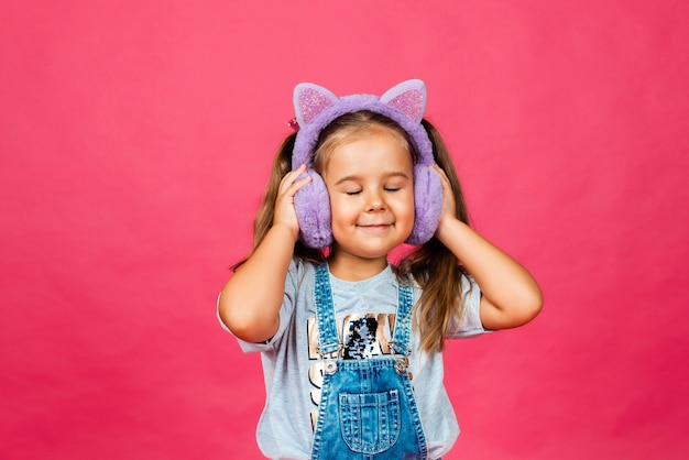 Cute smiling little girl having fun in fur headphones on a pink background.