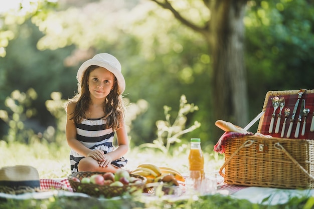 A cute smiling little girl is sitting on a grass and enjoying picnic day in the park.