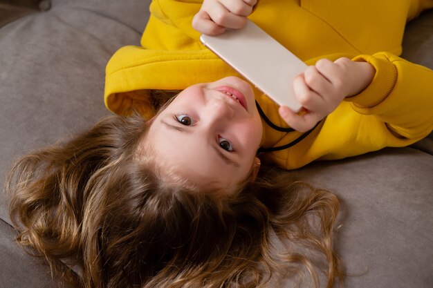 cute smiling little girl in yellow clothes is lying on her back on the bed and holding a smartphone