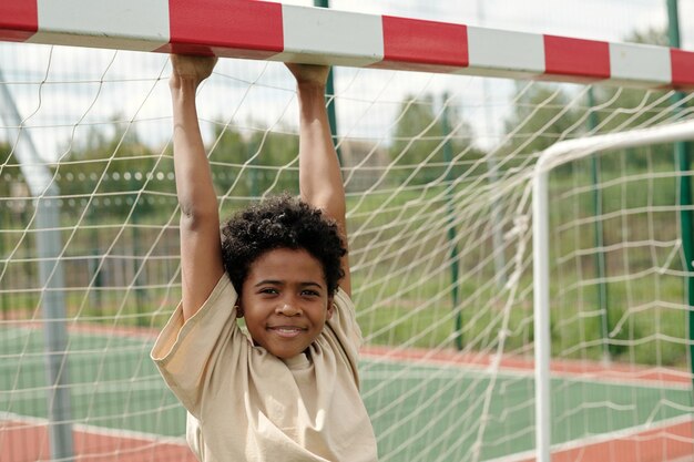 Cute smiling schoolboy in tshirt hanging on goal gate and looking at camera