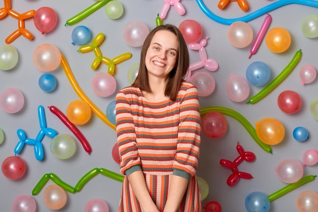 Cute smiling shy woman wearing striped dress standing against gray wall with balloons decoration looking at camera waiting for guests having joyful expression