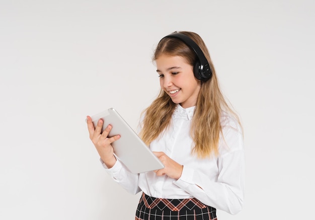 Cute smiling teen girl in school uniform with headphones and tablet in her hands isolated on white