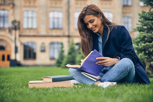 Милая улыбающаяся женщина сидит в одиночестве на траве возле университета во время подготовки к экзамену