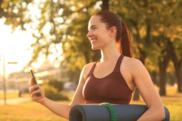 Cute smiling woman wearing tracksuit using smartphone and holding yoga mat at sunset in park.