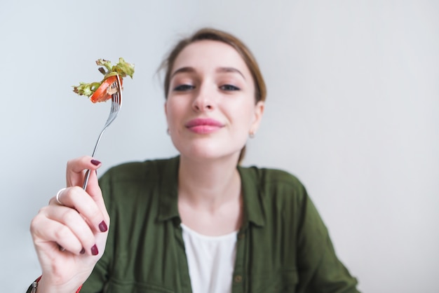 cute smiling woman with fork of salad in her hand is isolated on gray background.